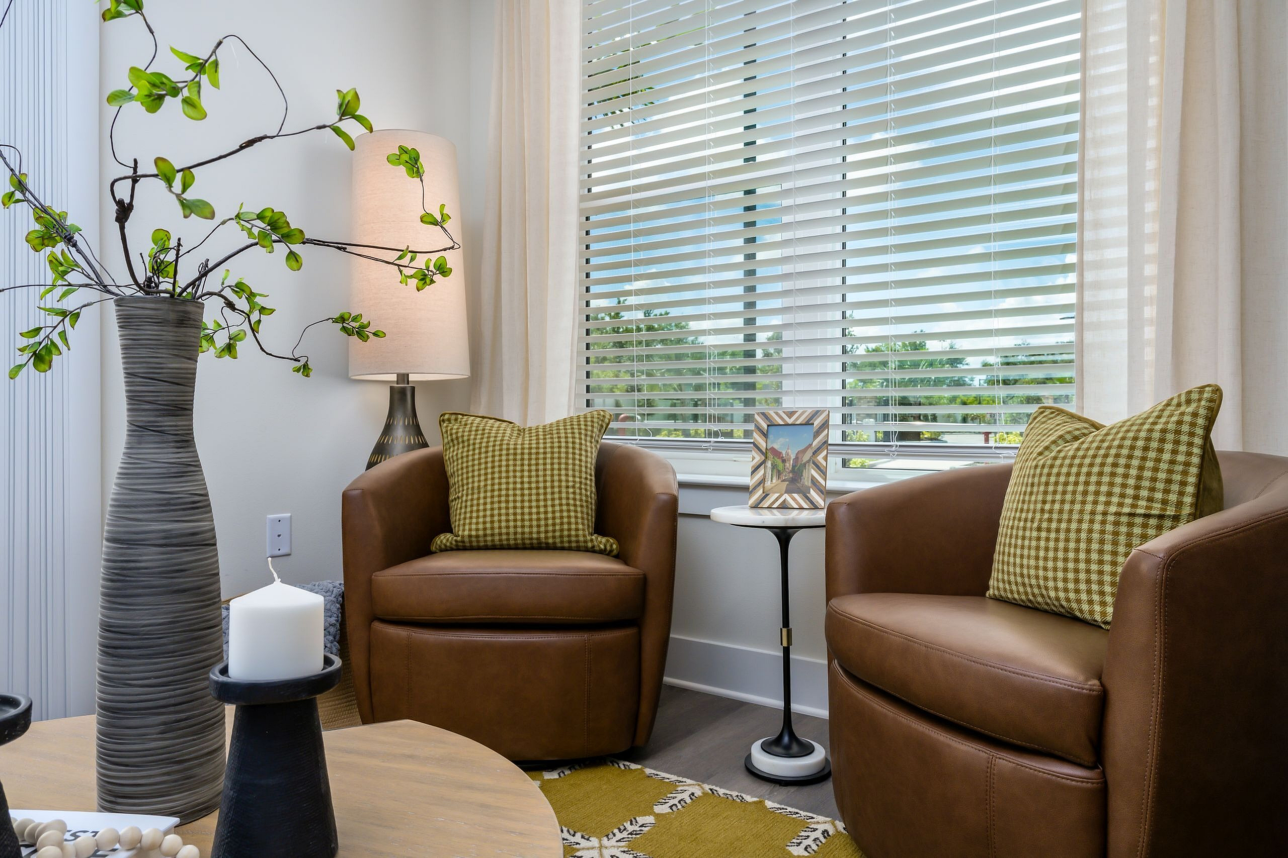 Cozy living room corner with elegant brown leather chairs, stylish greenery in a modern vase, and warm natural light streaming through blinds.