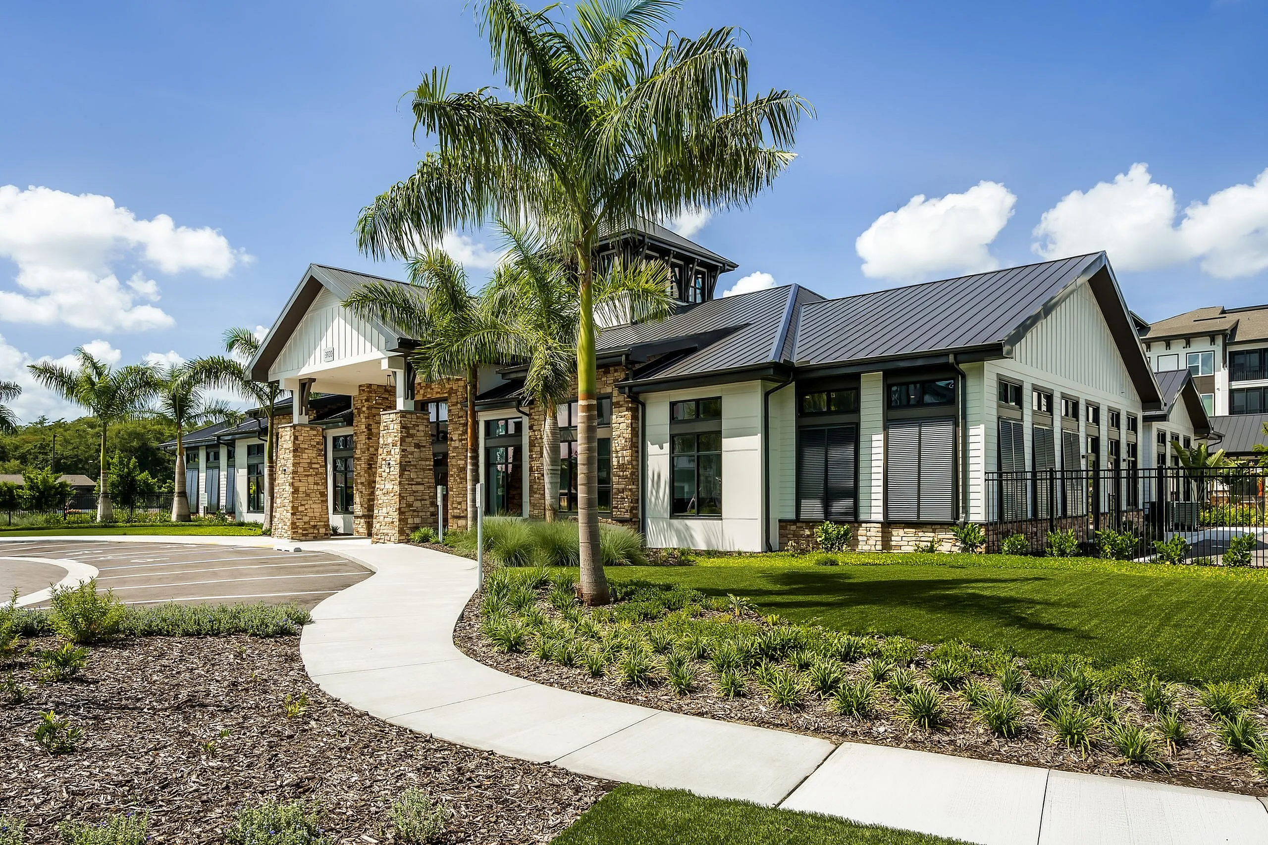 Luxurious modern clubhouse surrounded by lush landscaping and palm trees, featuring a sleek metal roof and stone columns.