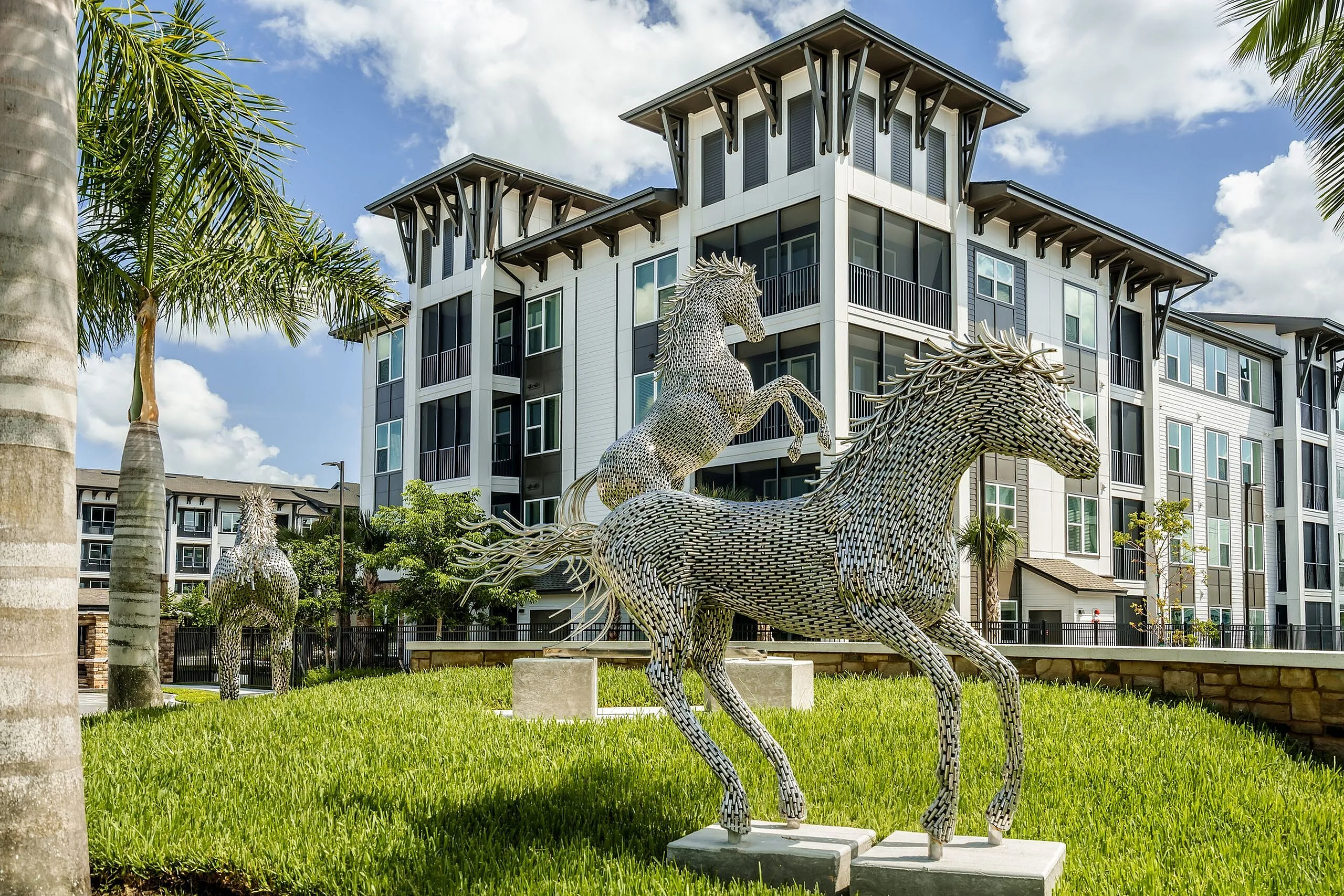 Contemporary apartment complex surrounded by lush greenery, featuring striking horse sculptures and modern architectural design.