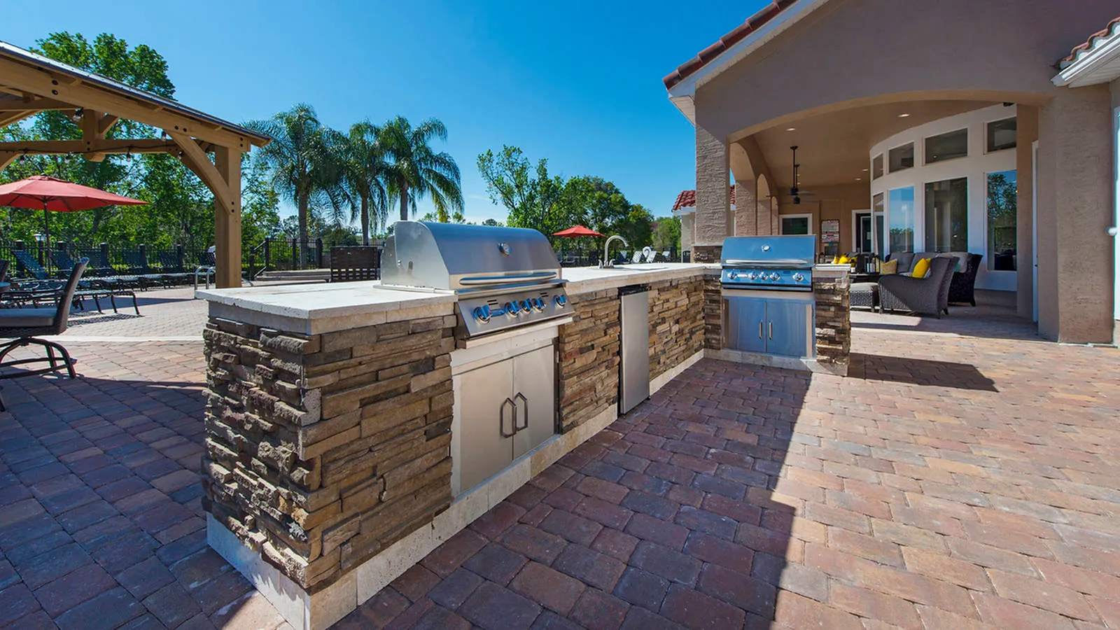 Outdoor grills on brick pavers. Trees and pool in background.