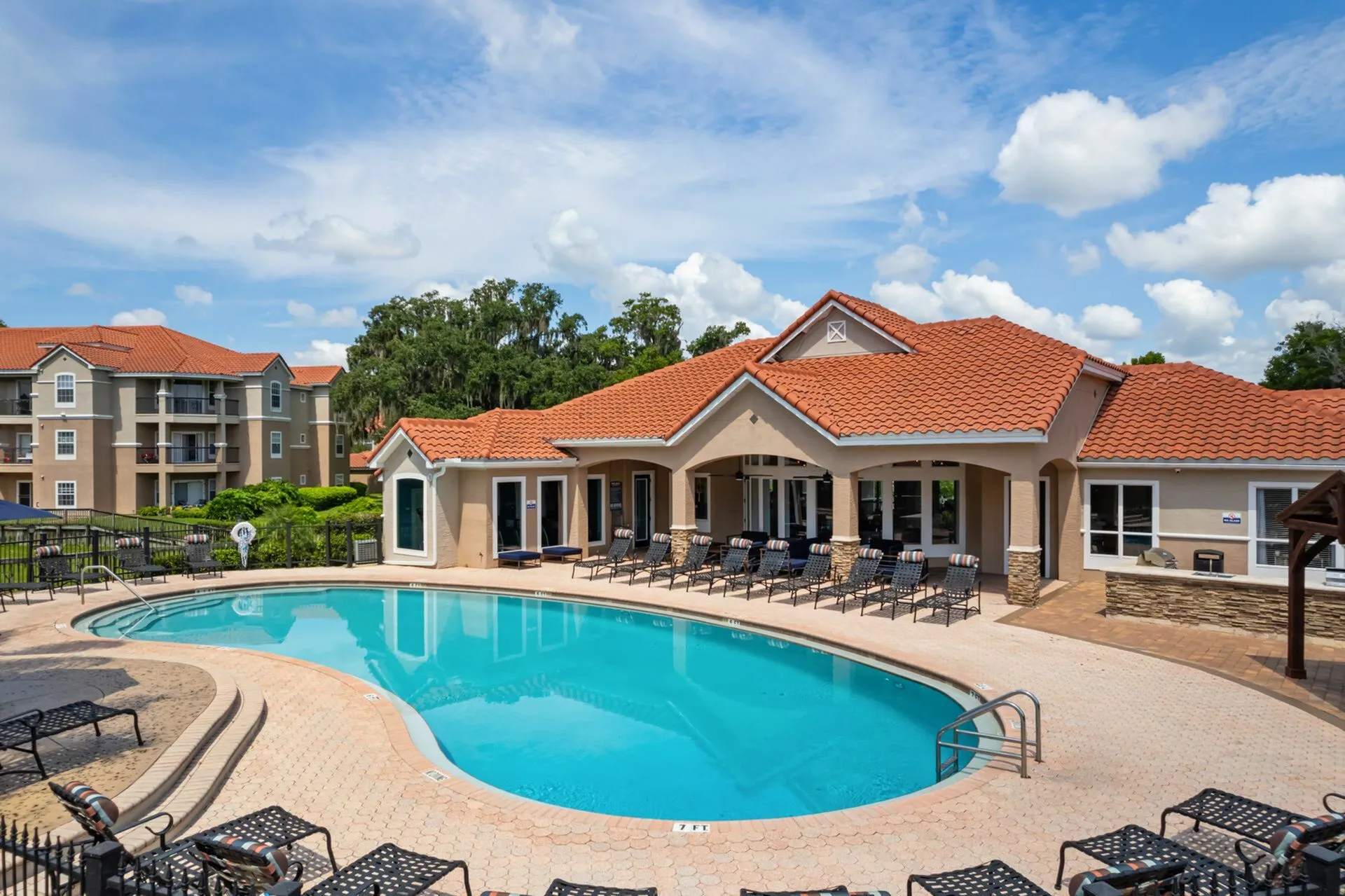 Luxurious apartment complex with a sparkling pool, inviting sundeck, and charming terracotta-roofed clubhouse under a bright blue sky.