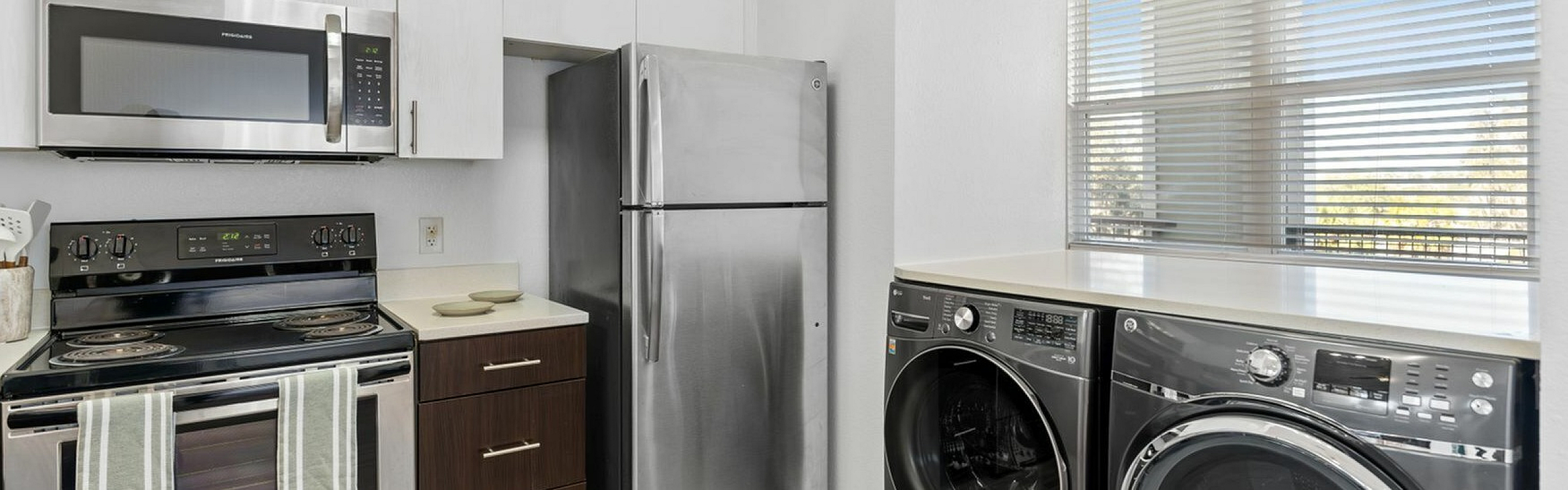 Modern kitchen with sleek stainless steel appliances, bright cabinetry, and an in-unit washer and dryer under a sunlit window.