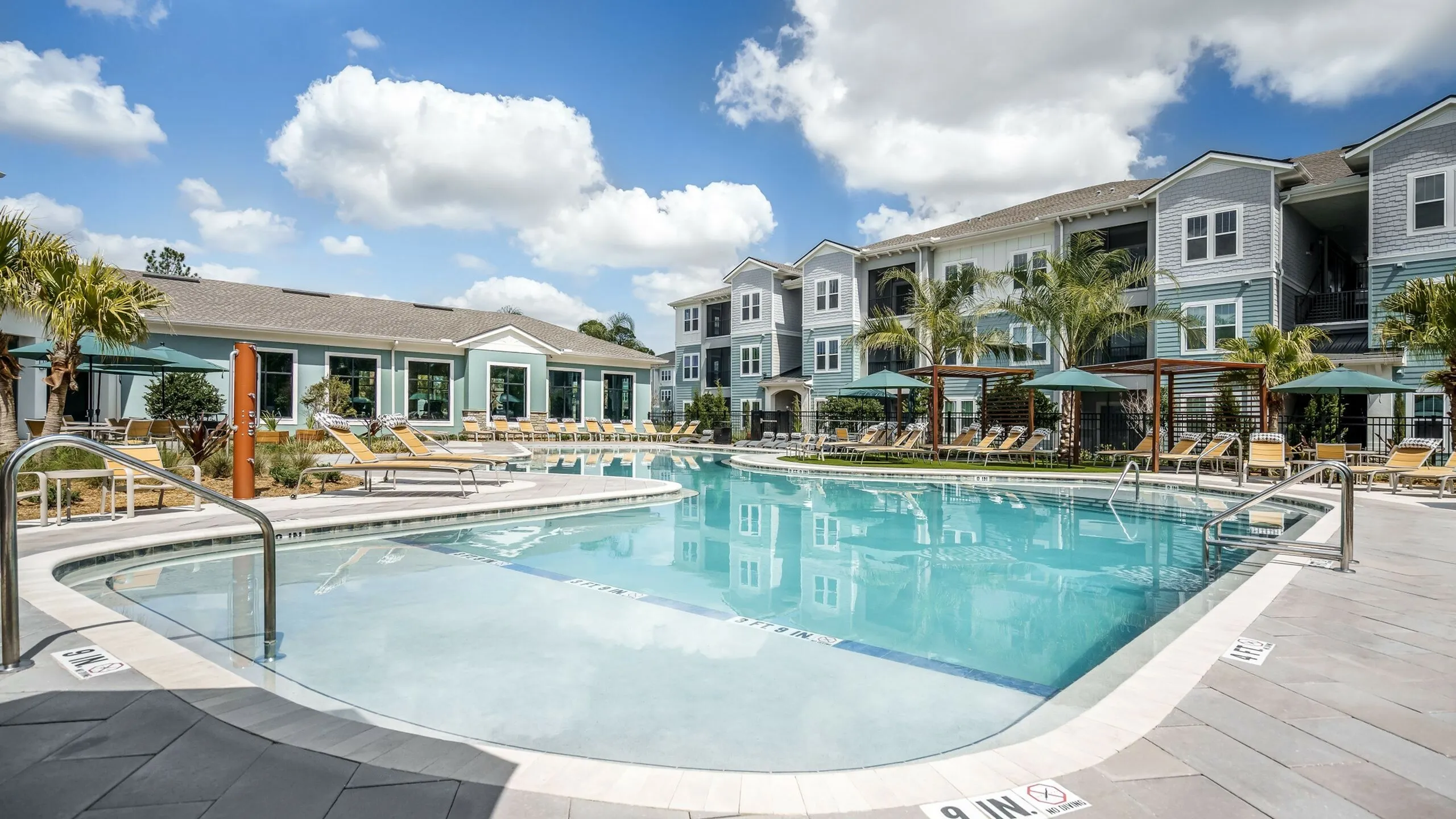 Luxurious apartment complex with a sparkling outdoor pool, sun loungers, and elegant landscaping under a bright blue sky.