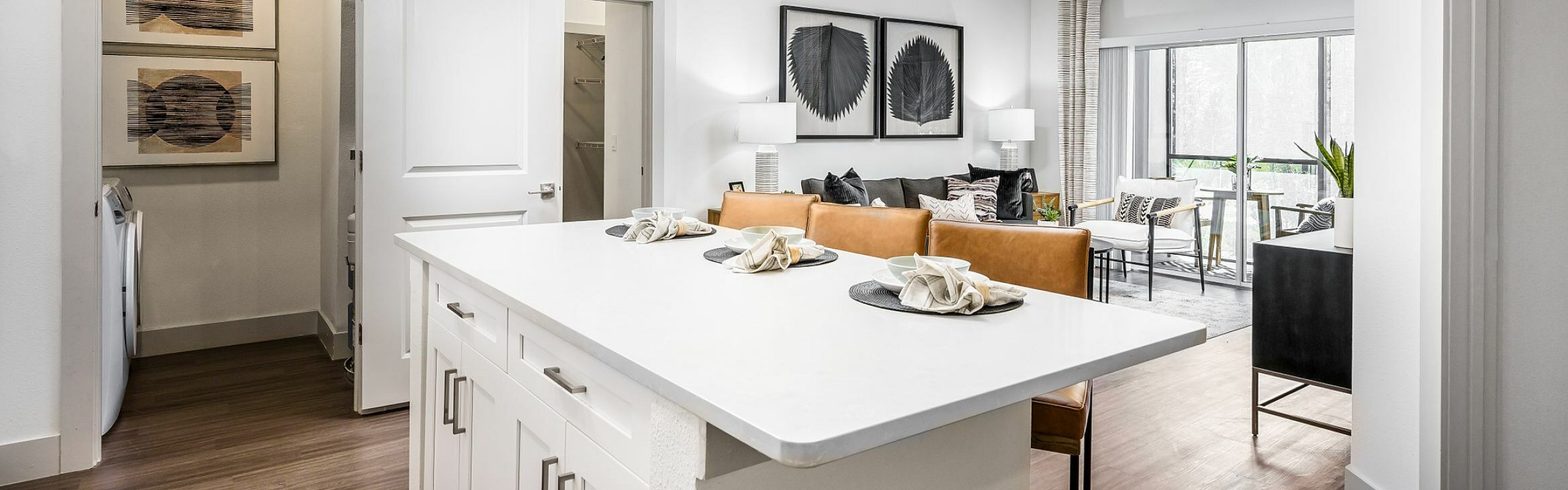 Modern kitchen and living area with sleek white cabinetry, elegant pendant lighting, and a cozy lounge space opening to a bright patio.