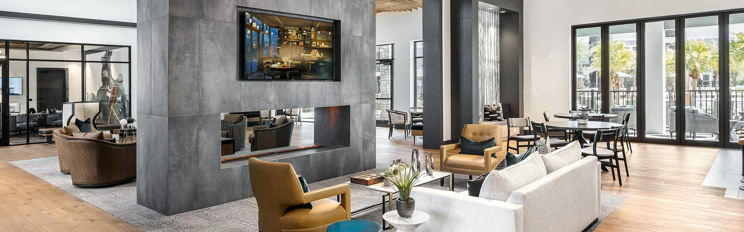 Contemporary apartment lounge with elegant decor, featuring a chic seating area, modern fireplace, and large windows overlooking a sunlit terrace with palm trees.