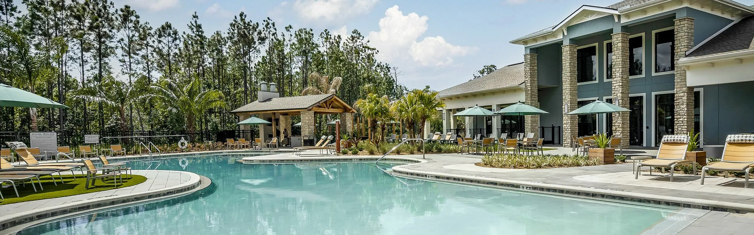 Luxurious apartment community with a sparkling pool, sun loungers, and lush landscaping under a bright blue sky.