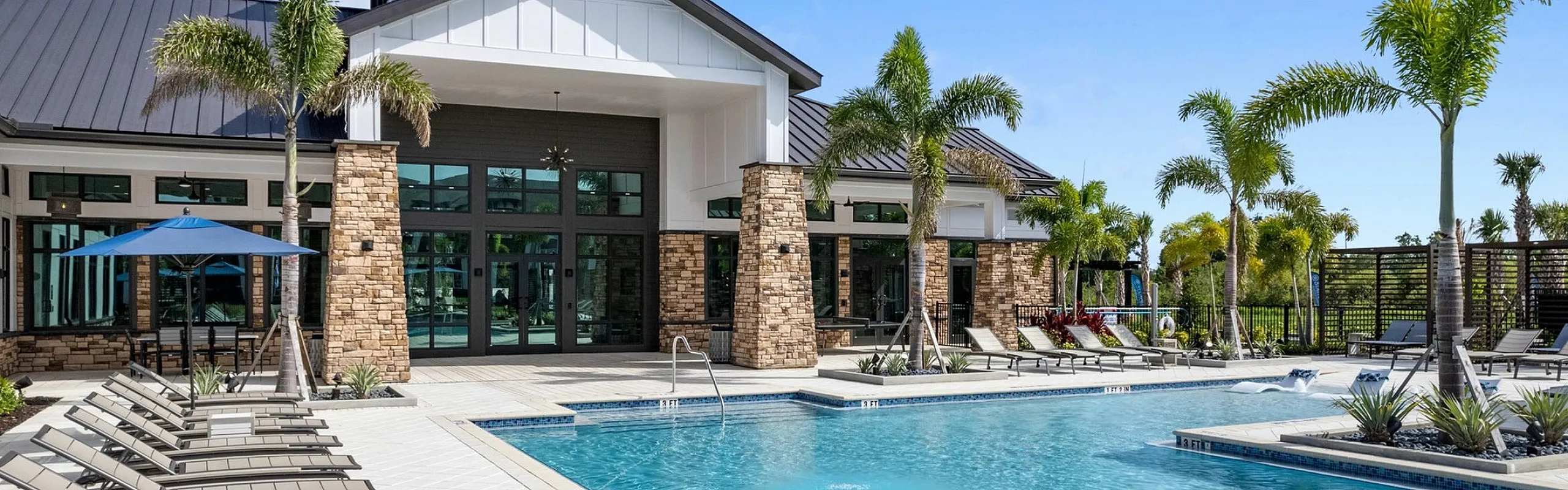 Luxurious outdoor pool area with sun loungers and elegant clubhouse surrounded by palm trees under a clear blue sky.