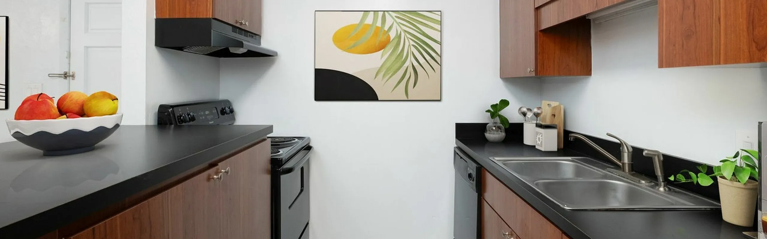 Modern kitchen featuring sleek black countertops, warm wooden cabinets, and stylish decor with vibrant artwork and fresh fruit bowl.