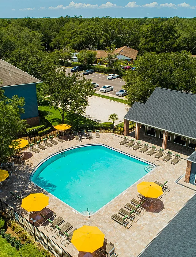 Aerial view of a luxury apartment complex featuring a pristine swimming pool surrounded by yellow umbrellas, lush greenery, and ample lounge chairs, creating a resort-style oasis.