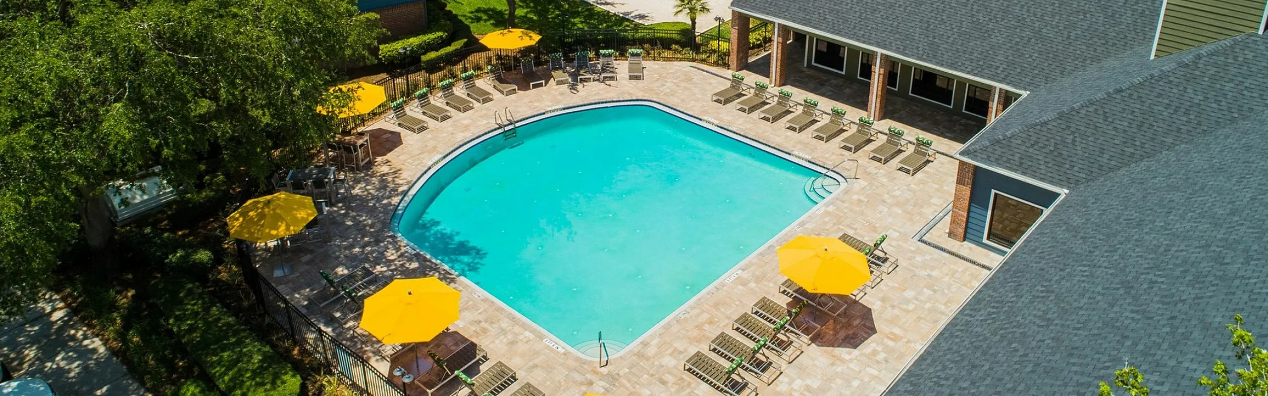 Aerial view of a luxury apartment complex with a sparkling blue swimming pool, surrounded by sun loungers and vibrant yellow umbrellas, amidst lush green trees.