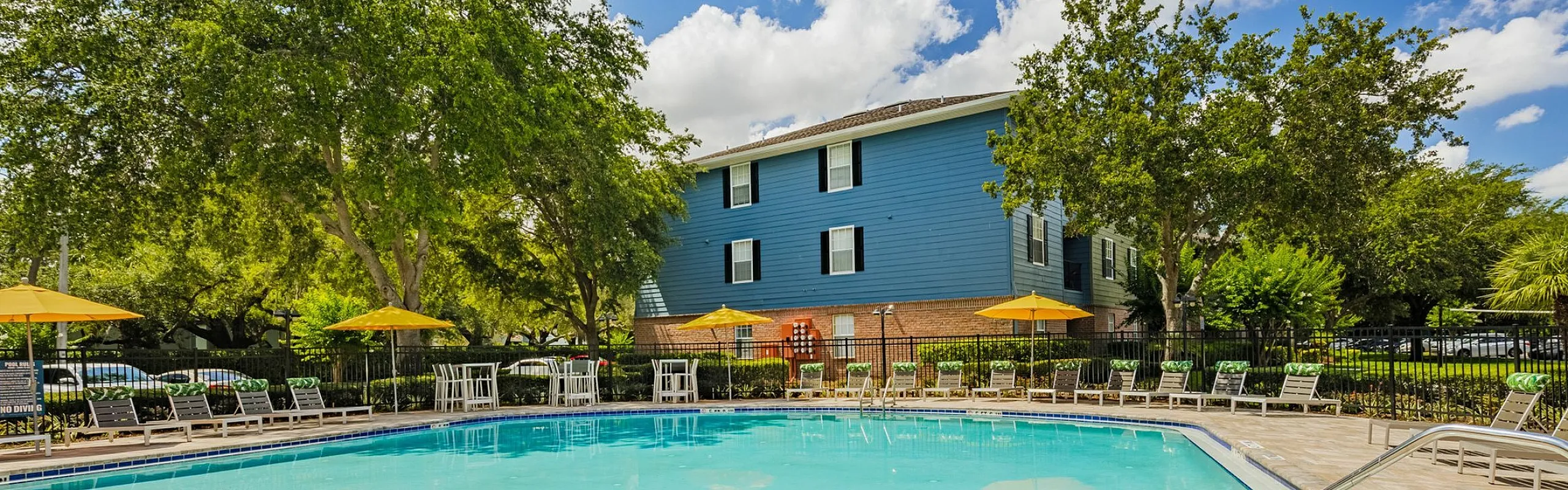 Inviting outdoor pool area with sunny lounge chairs and vibrant yellow umbrellas, surrounded by lush green trees and a charming blue apartment building under a bright blue sky.
