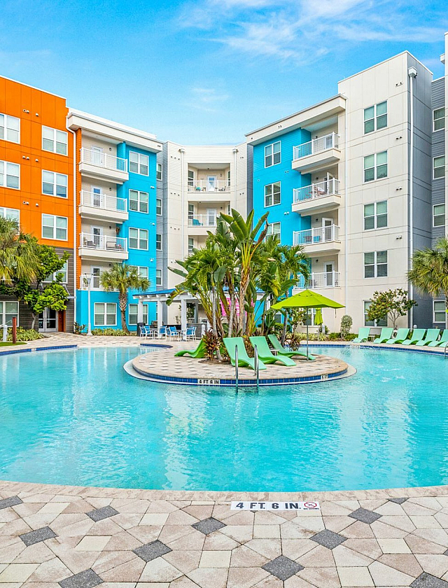 Vibrant apartment complex with colorful exterior, palm trees, and a stunning curved pool surrounded by sun loungers, perfect for relaxation and leisure.