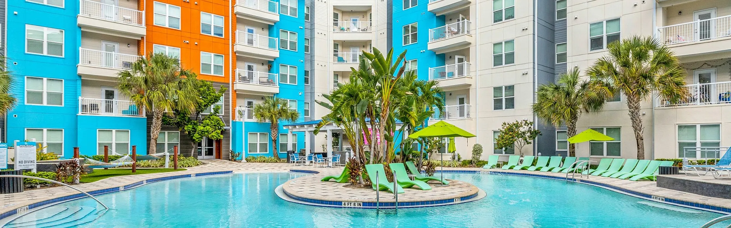 Vibrant apartment complex with colorful facades, featuring a luxurious pool surrounded by palm trees and sun loungers.