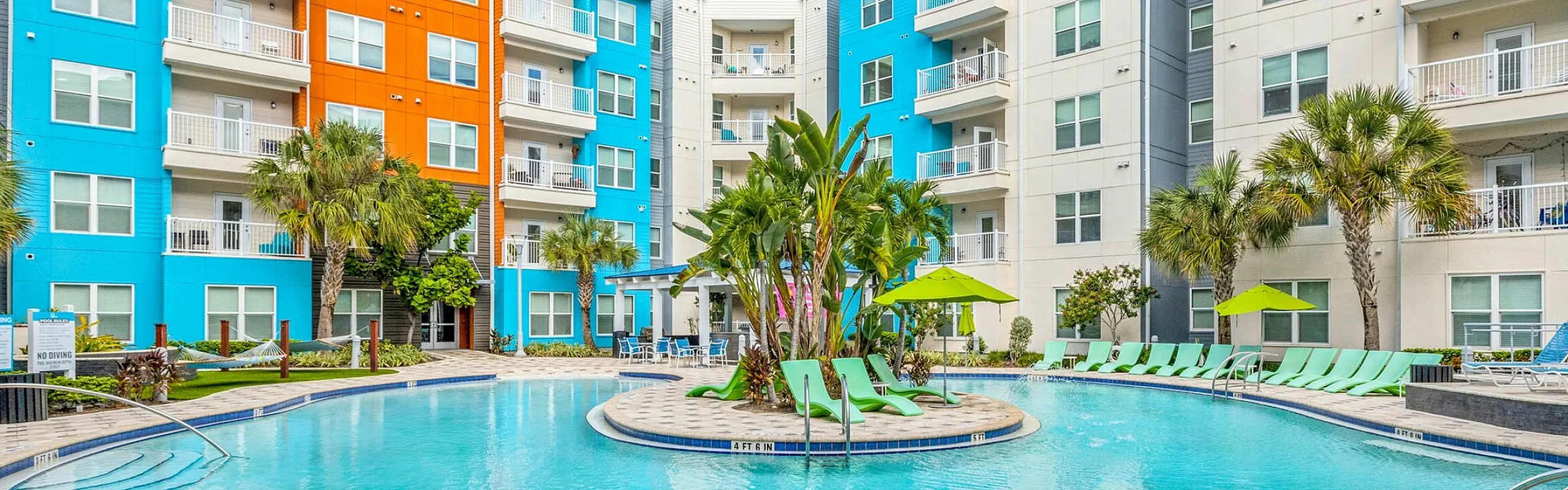 Vibrant apartment complex featuring colorful facades, a stunning pool with lush palm trees, and relaxing lounge areas.