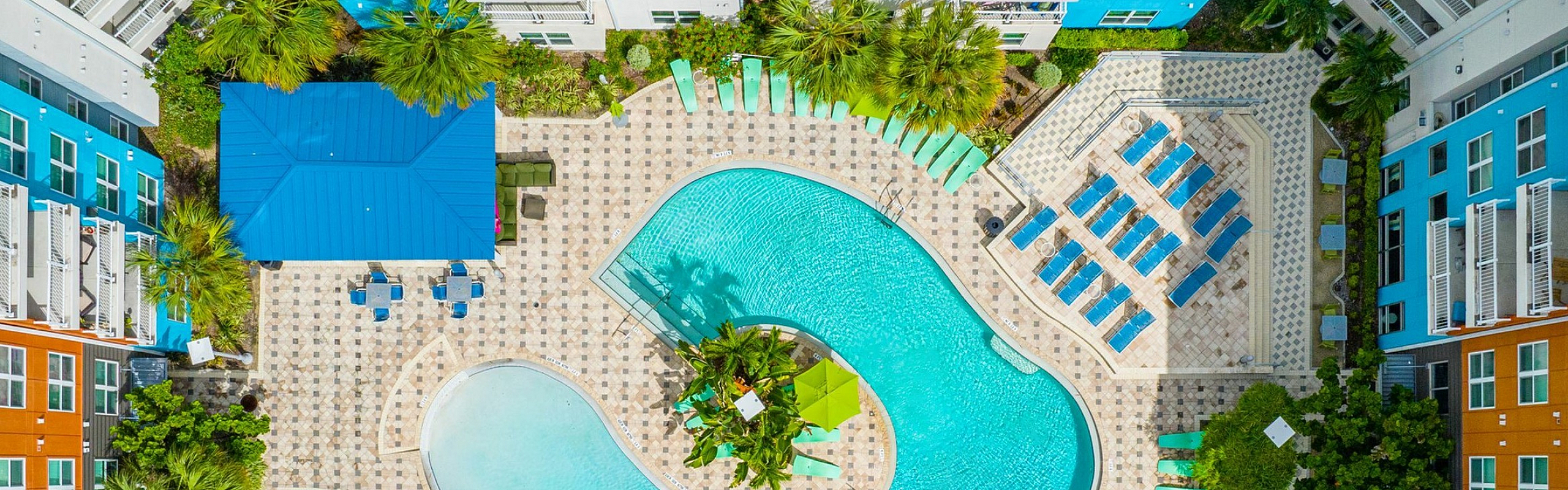 Vibrant aerial view of modern apartment complex with expansive pool area, lush greenery, and colorful sun loungers, creating a resort-style ambiance.