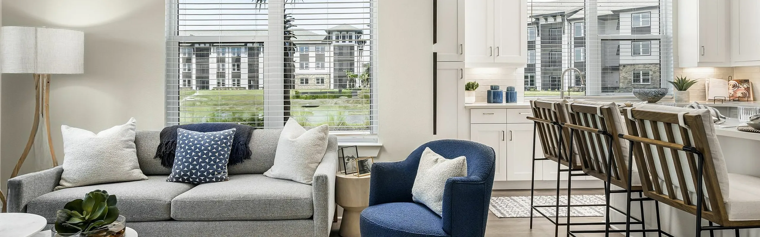 Bright, modern living room and kitchen space with stylish decor, featuring a cozy grey sofa, a chic blue chair, and a breakfast bar with elegant stools, overlooking a lush green outdoor view.