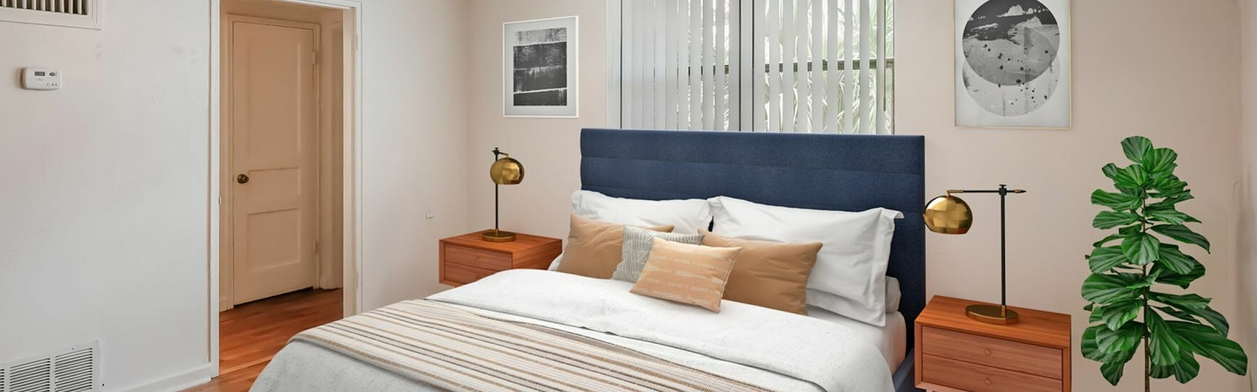 Inviting bedroom featuring a cozy bed with a blue headboard, elegant nightstands with gold lamps, and a lush potted plant, illuminated by natural light.