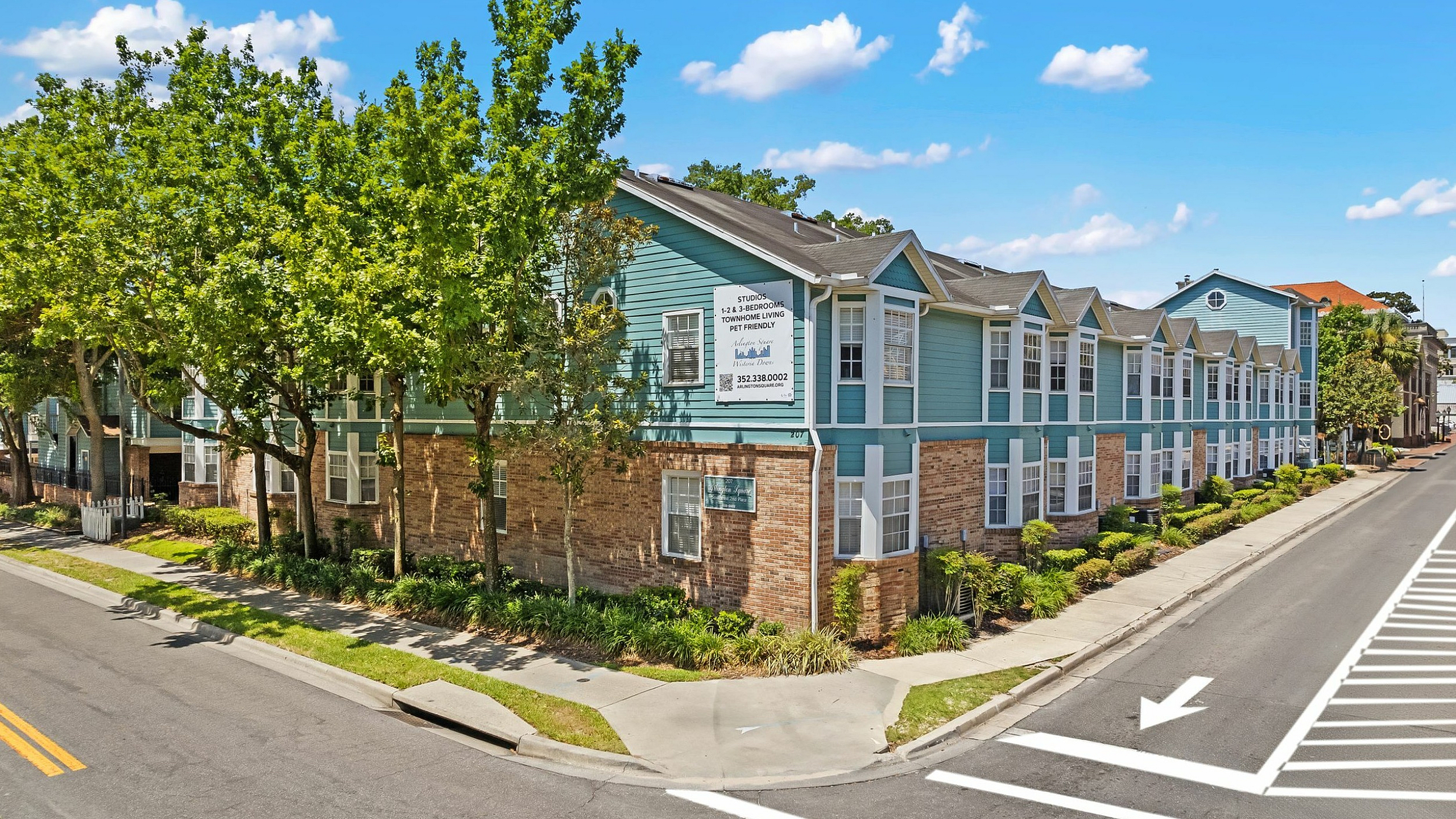 Charming apartment complex with modern blue and brick exterior, surrounded by lush greenery on a sunny day.