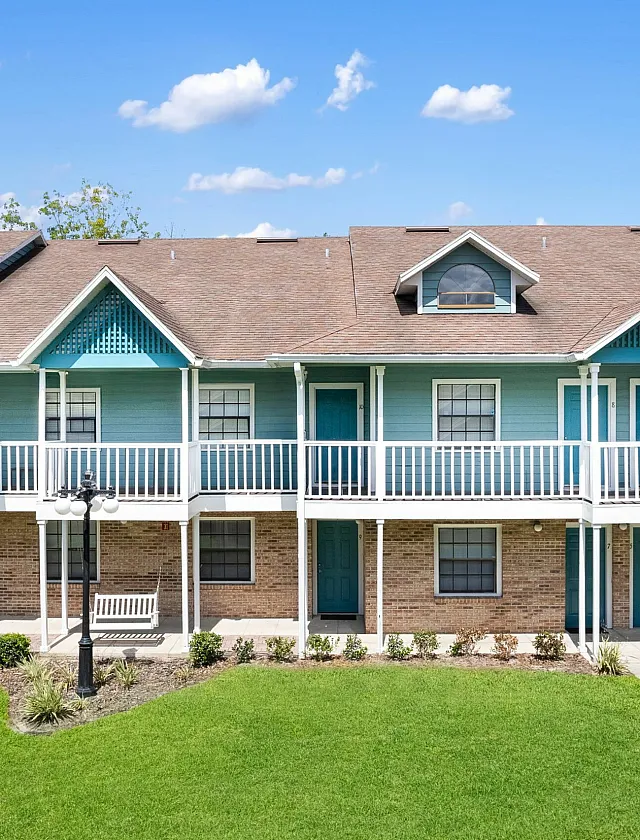 Charming apartment building with a vibrant blue exterior, offering spacious balconies and lush green surroundings under a bright, sunny sky.