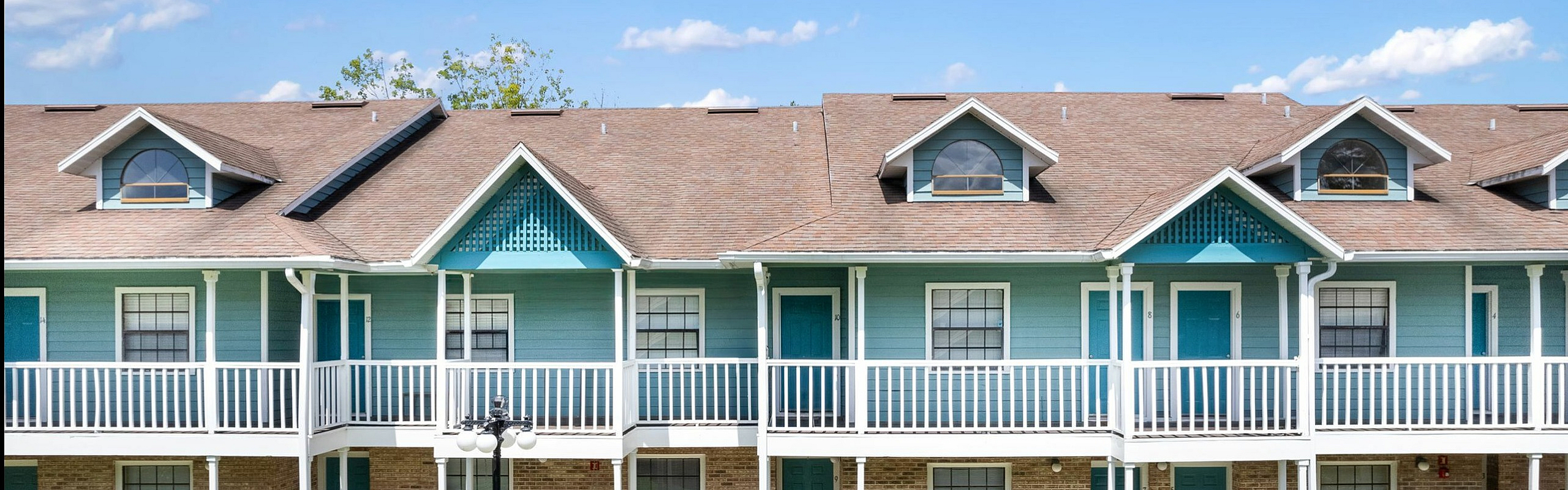 Charming two-story apartment complex with a turquoise and brick facade, featuring spacious balconies and lush green surroundings under a bright blue sky.