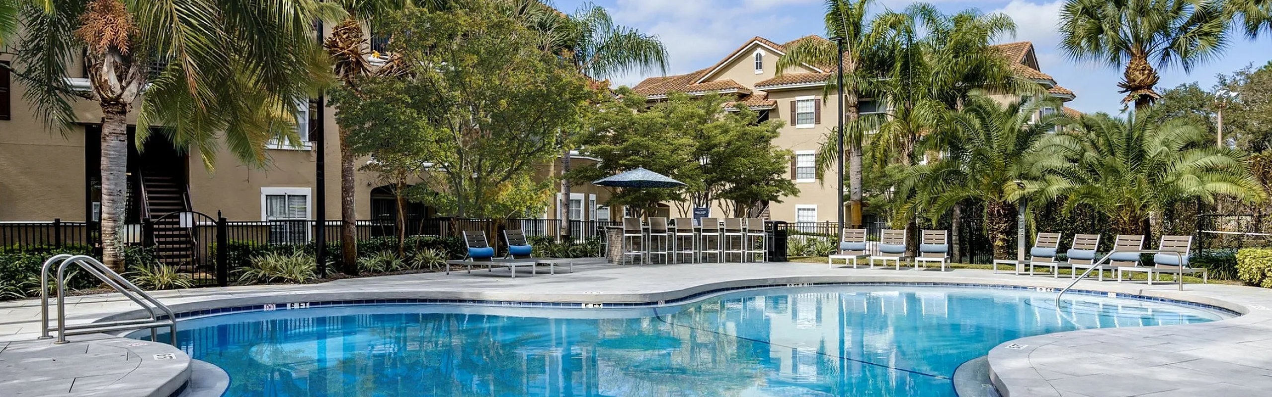 Resort-style swimming pool surrounded by lush palm trees and elegant apartment buildings under a bright blue sky.