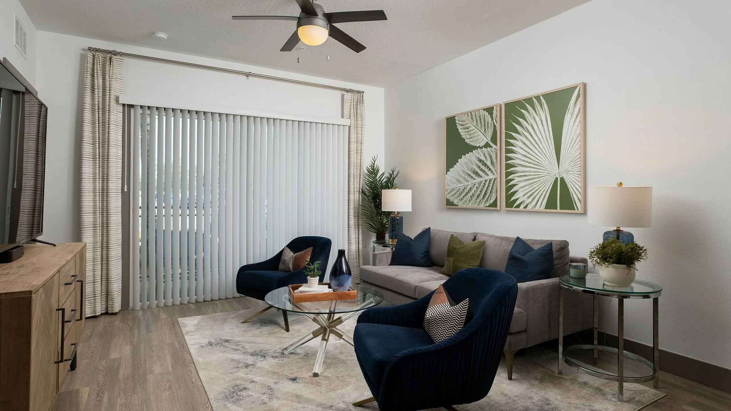 Elegant living room with modern decor, featuring stylish navy chairs, a sleek glass coffee table, and vibrant green artwork, under a sophisticated ceiling fan.