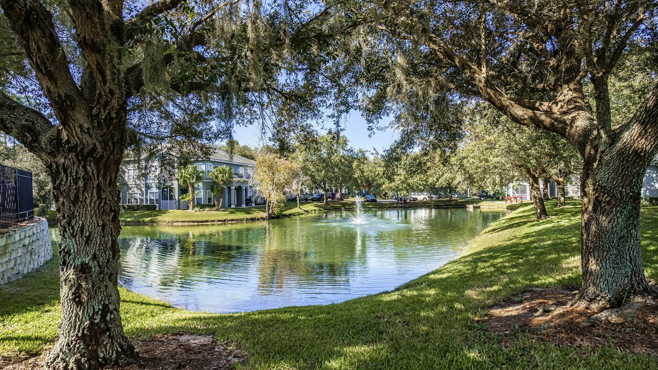 Tranquil apartment view featuring lush trees, a serene pond with a fountain, and a charming building in the background.
