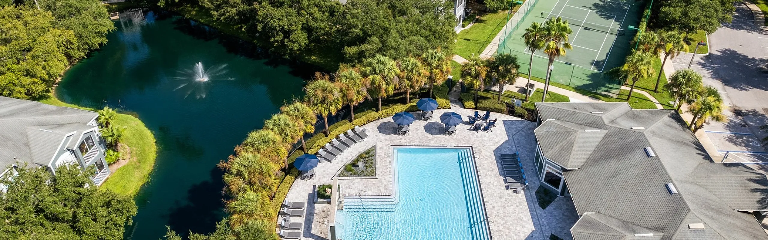 Aerial view of a luxurious apartment community featuring a sparkling pool with lounging chairs, a serene pond with a fountain, and a tennis court surrounded by lush greenery.