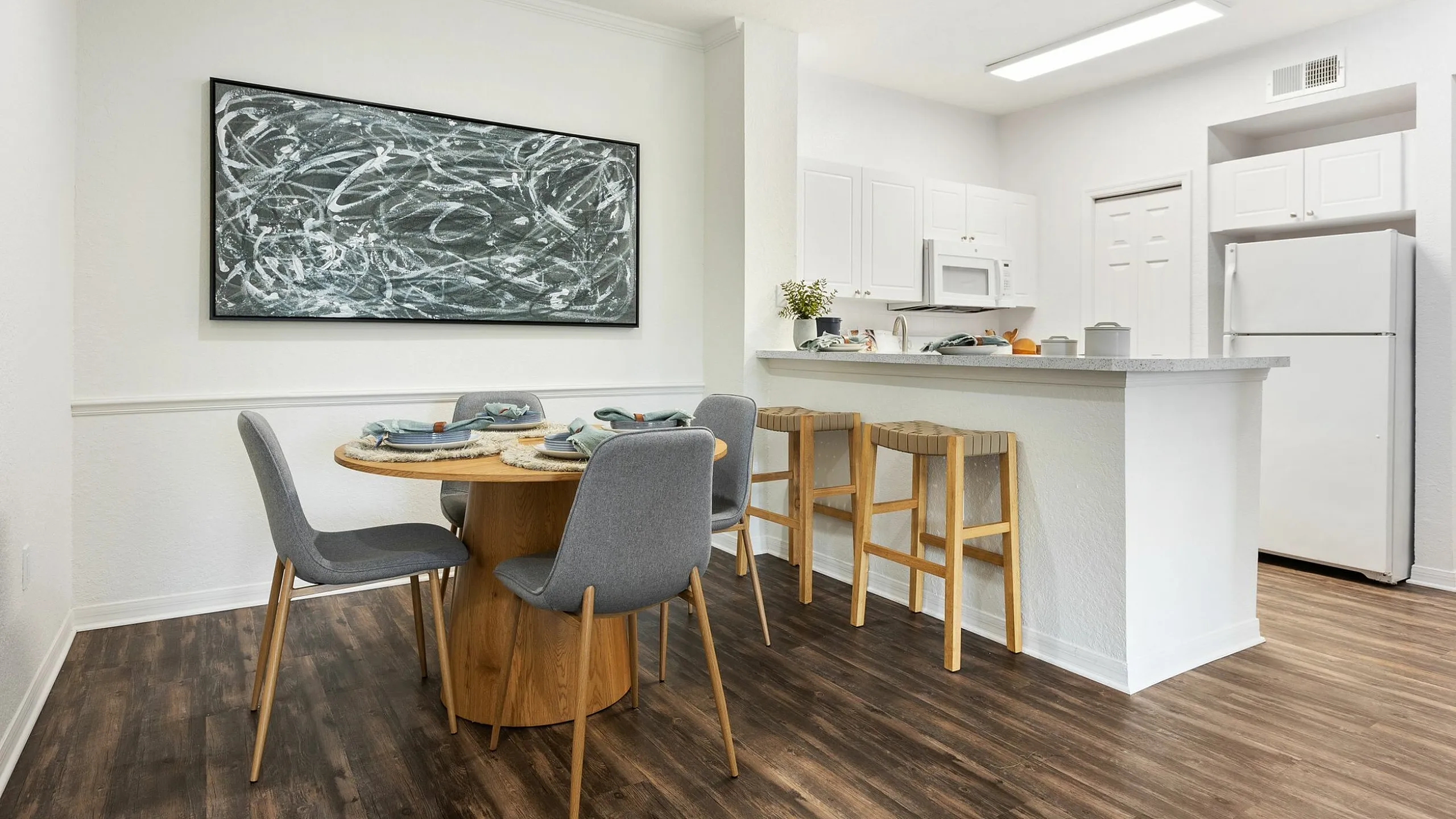 Chic dining area with modern art, cozy wooden round table set for dinner, and open-concept kitchen with sleek white cabinetry and a breakfast bar.