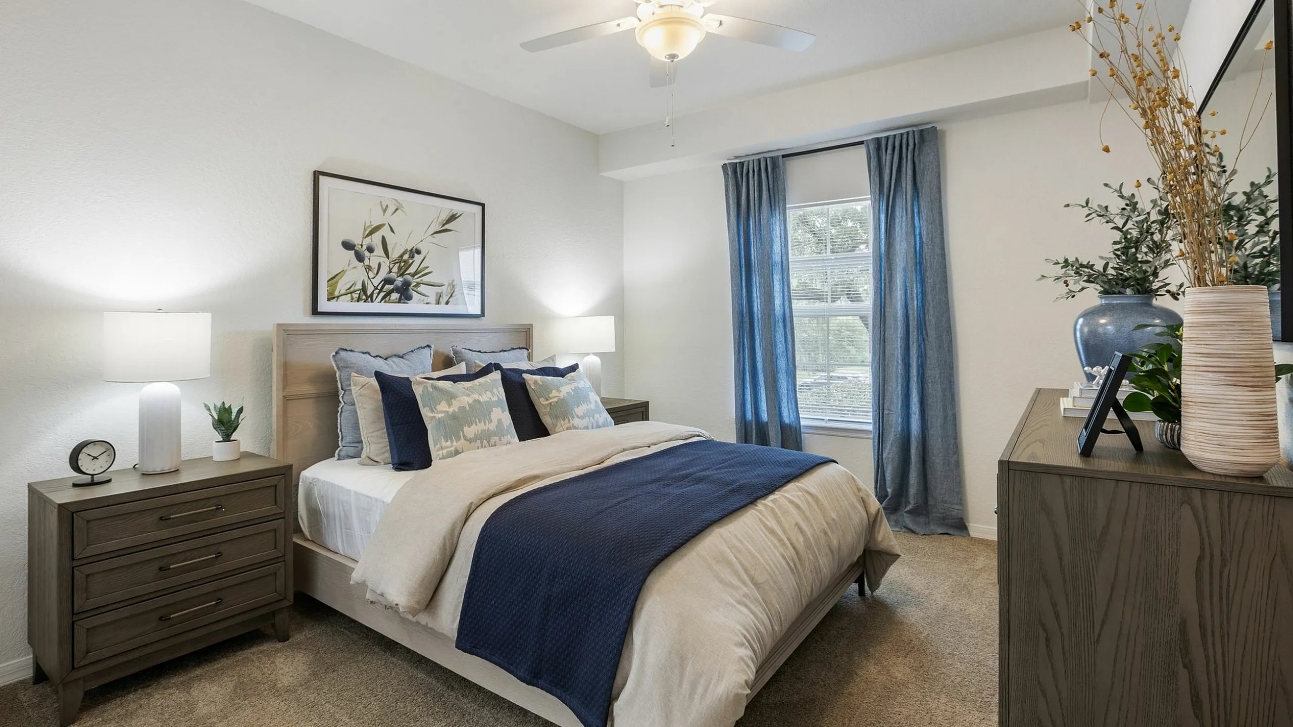 Inviting bedroom with elegant decor, featuring a comfortable bed with plush pillows, modern nightstands with stylish lamps, and abundant natural light through large windows with blue curtains.