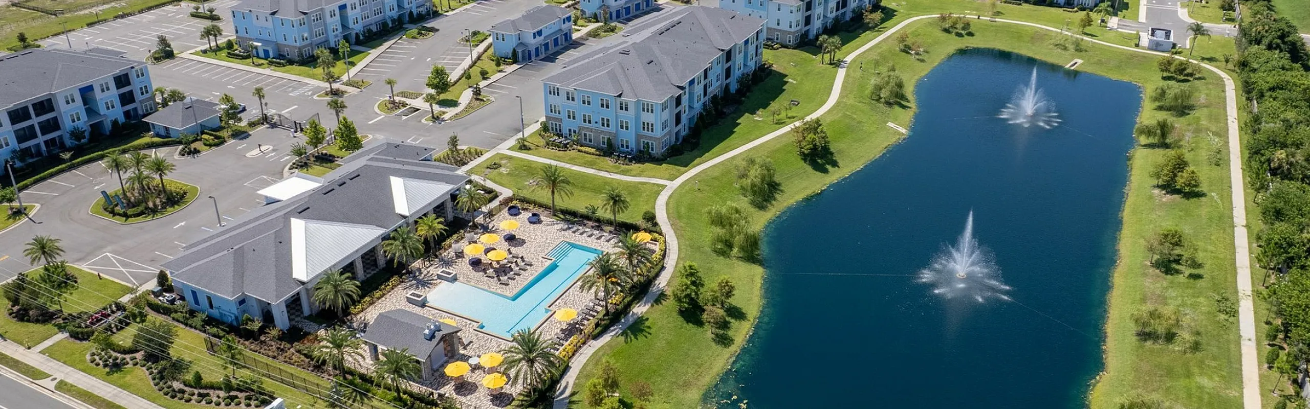 Aerial view of a serene apartment community featuring multiple buildings, a sparkling blue pool with sun umbrellas, and a picturesque pond with fountains surrounded by lush green landscape.