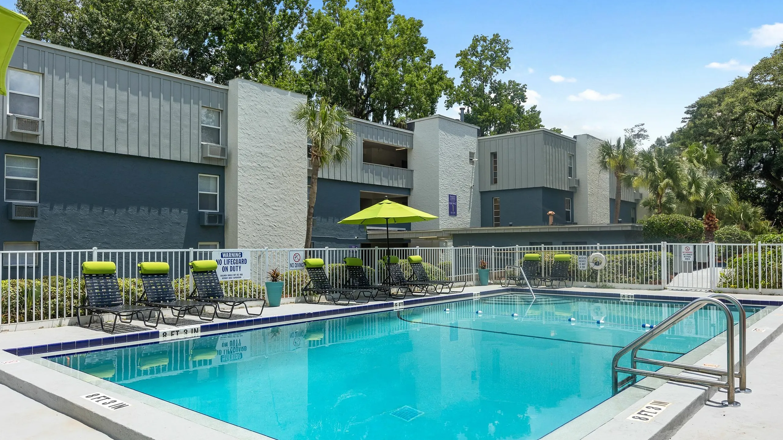 Modern apartment complex featuring a sparkling outdoor pool with bright green lounge chairs and umbrellas, surrounded by lush trees and stylish buildings.