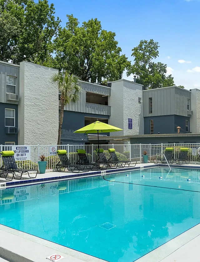 Inviting residential outdoor pool area surrounded by modern buildings and lush greenery, featuring stylish loungers and vibrant umbrellas.