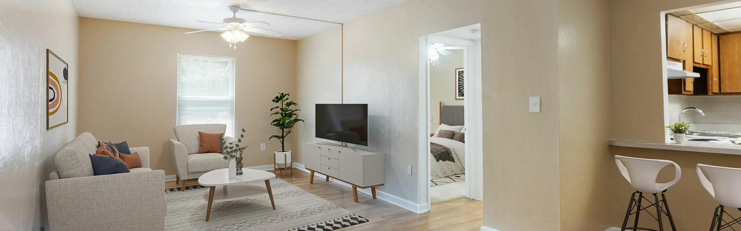 Charming living area with warm beige tones, featuring a cozy sofa set, stylish coffee table, and modern entertainment console; open layout connects to a quaint kitchen with elegant bar stools.