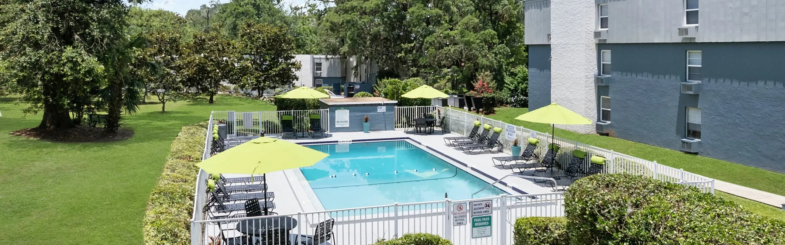 Inviting apartment complex with a pristine swimming pool, lounge chairs, and vibrant green umbrellas, surrounded by lush greenery.