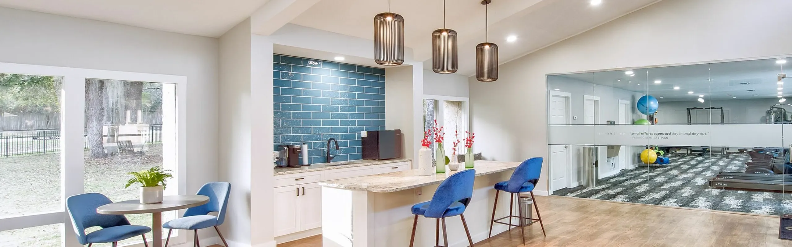 Bright open-concept lounge with modern kitchen island, stylish pendant lighting, and a view of the inviting fitness room.