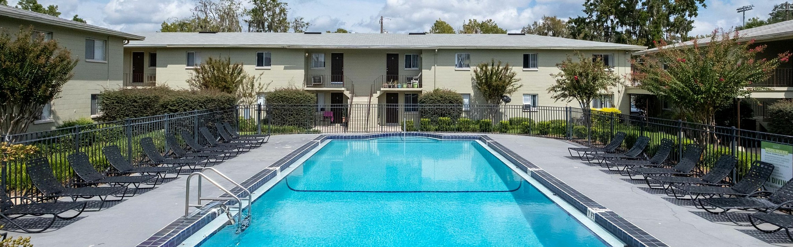 Inviting apartment complex showcasing a pristine swimming pool with ample lounge chairs, surrounded by lush landscaping and charming two-story buildings under a bright blue sky.