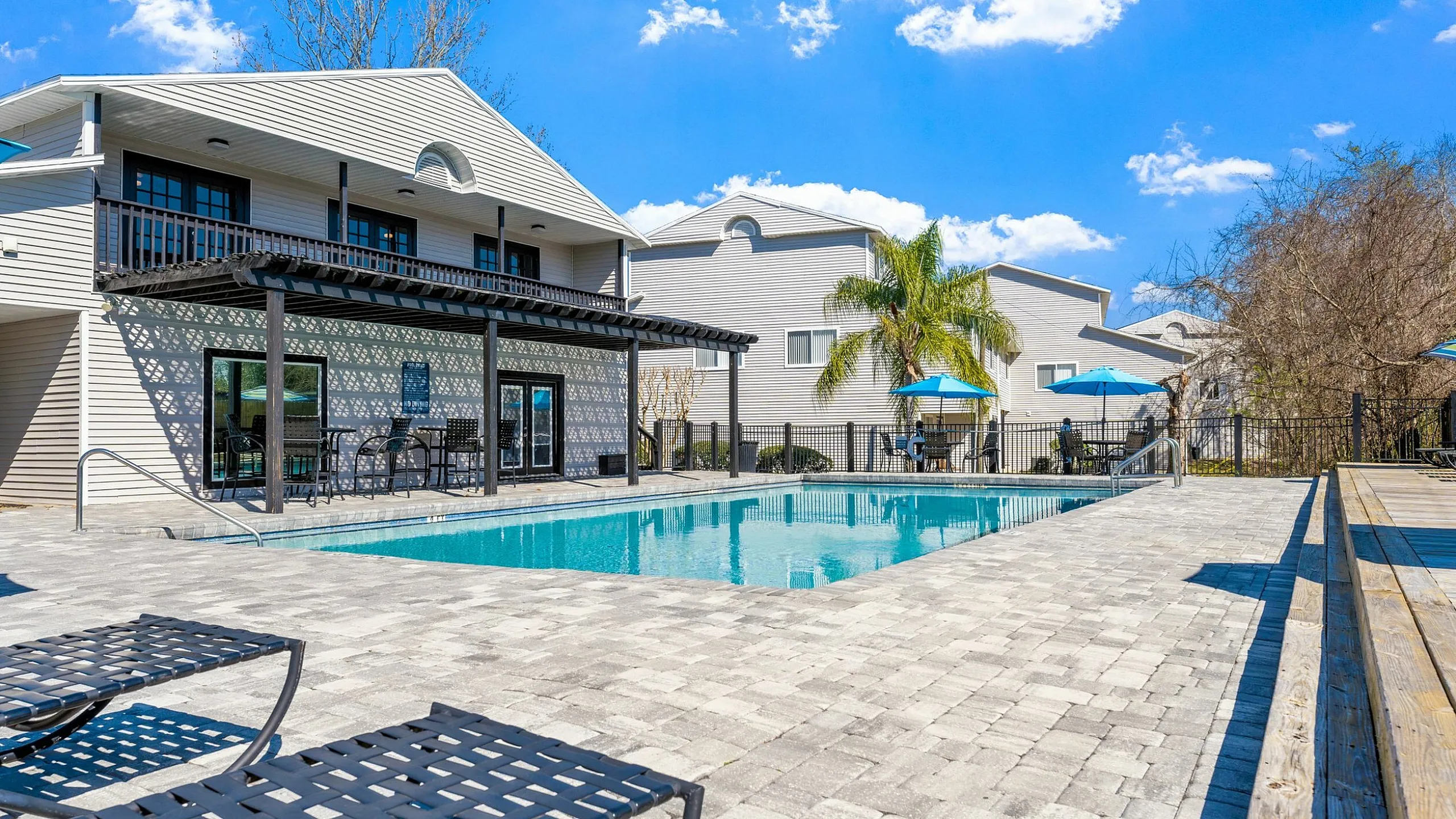 Charming apartment complex with a sparkling swimming pool, surrounded by sun loungers, modern architecture, and lush palm trees under a clear blue sky.