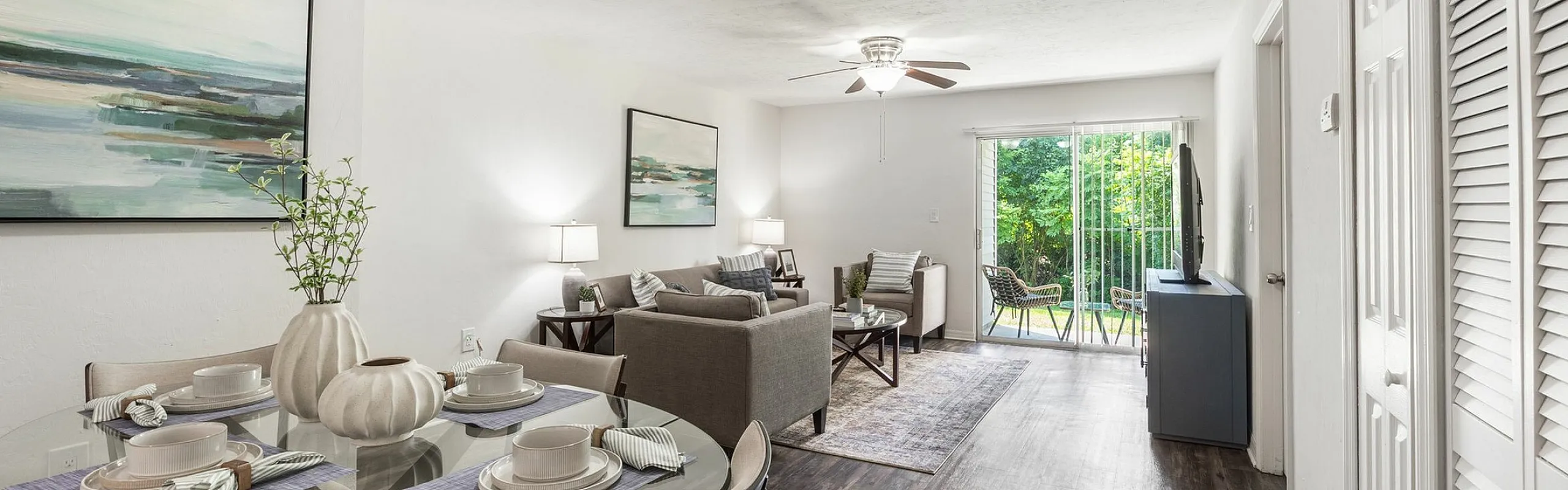 Elegant open-concept living and dining room with modern decor, glass dining table, cozy seating, and large glass doors leading to a lush green balcony view.