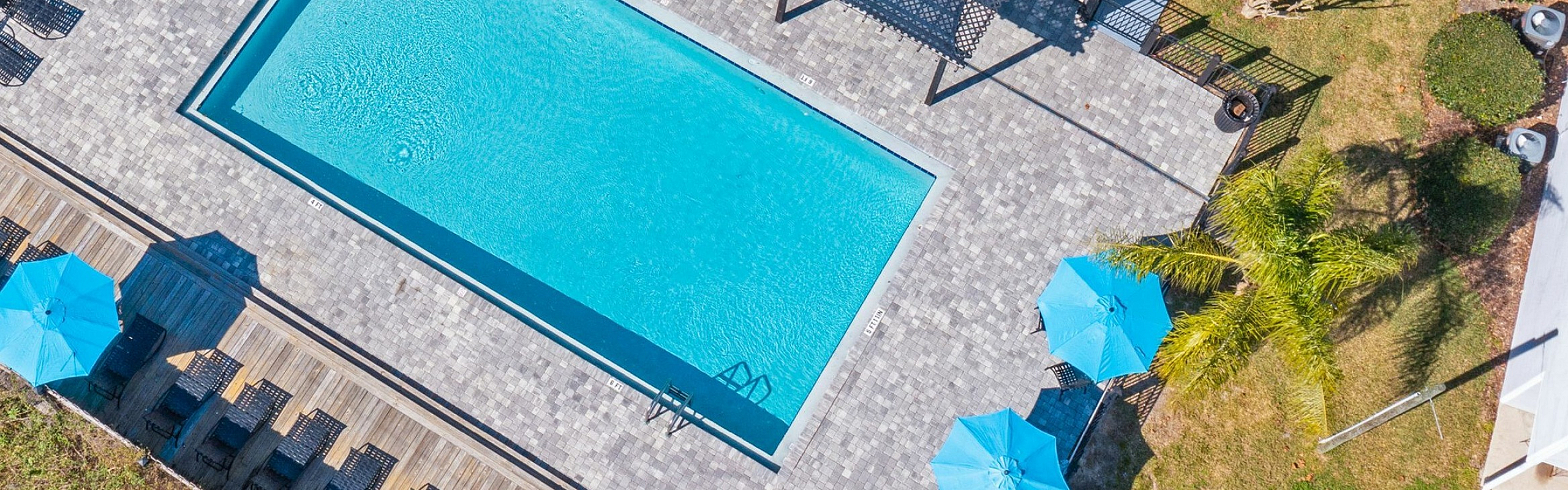 Aerial view of a modern apartment complex featuring a sparkling blue swimming pool, elegant deck area, and cozy seating with blue umbrellas, perfect for relaxation and enjoyment.