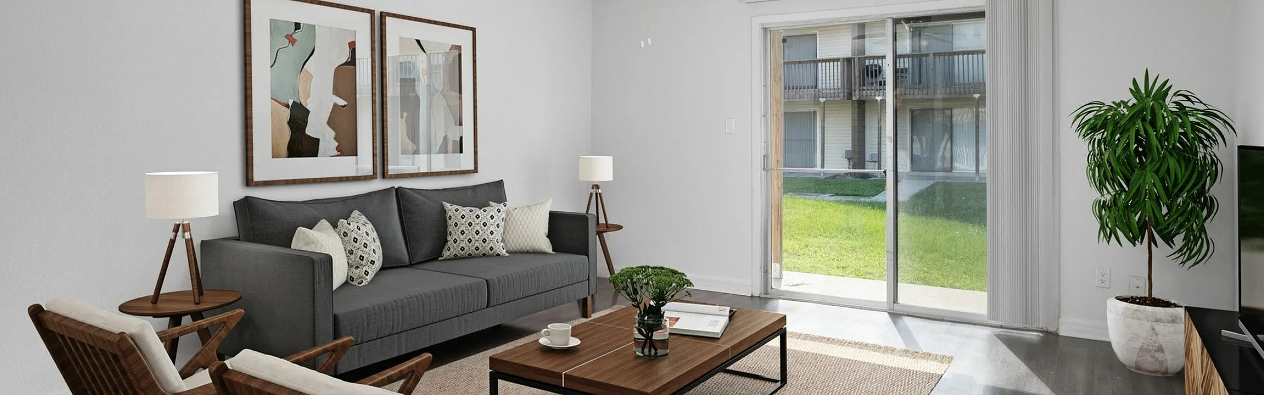 Bright and modern living room with sleek wooden flooring, cozy gray sofa, and stylish decor. Features large sliding glass doors opening to a lush green view.