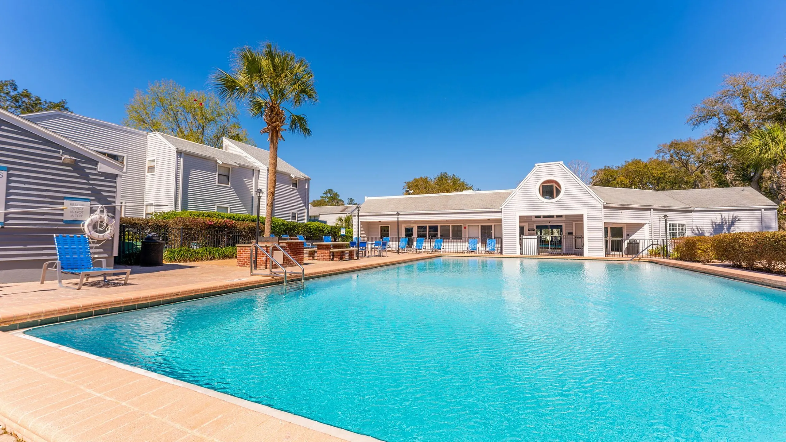 Inviting apartment complex featuring a sparkling outdoor swimming pool with lounge chairs, surrounded by modern architecture against a clear blue sky.