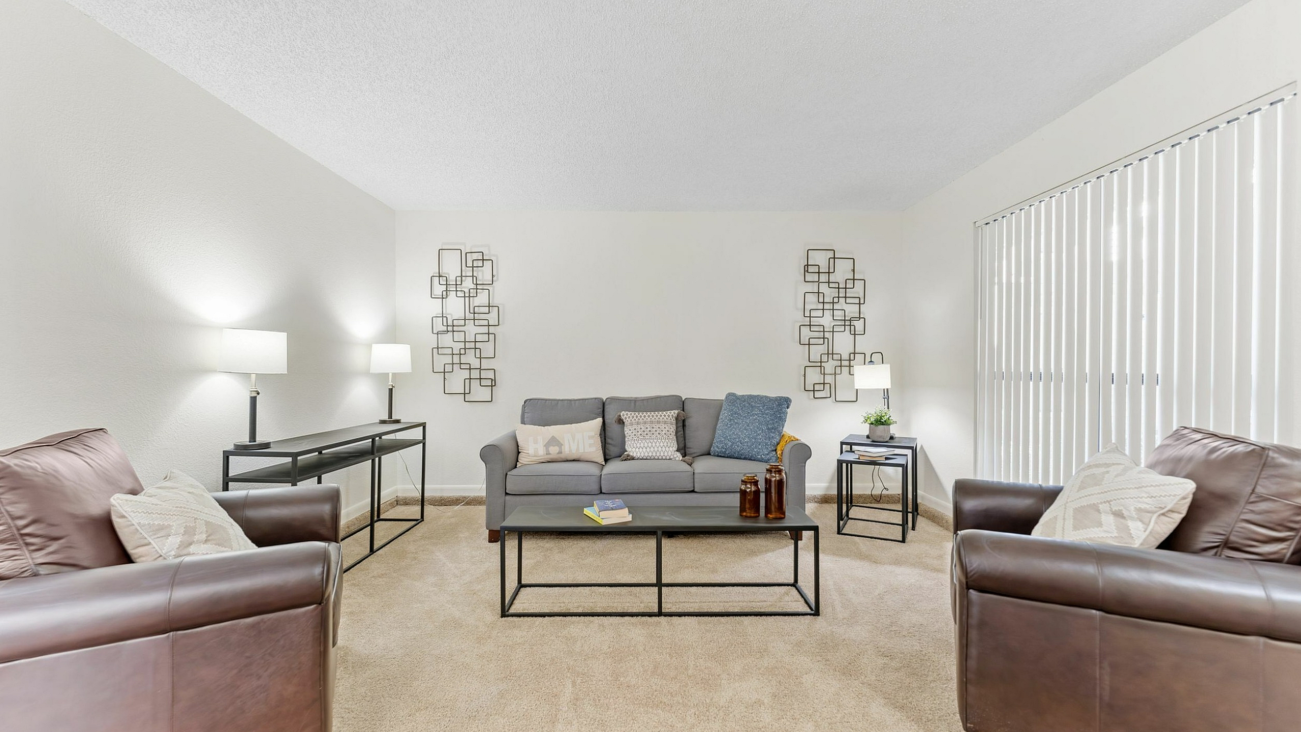 Inviting living room with cozy beige carpet, elegant leather sofas, and a chic gray couch. Abundant natural light pours in through vertical blinds, highlighting stylish geometric wall art and modern table lamps.