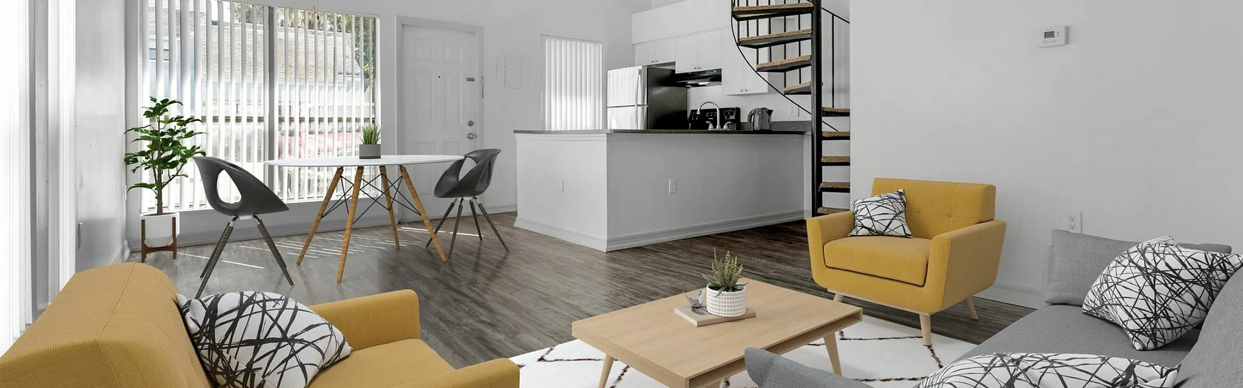 Modern open-plan apartment with stylish yellow and gray seating, elegant spiral staircase, and chic kitchen area under natural light.