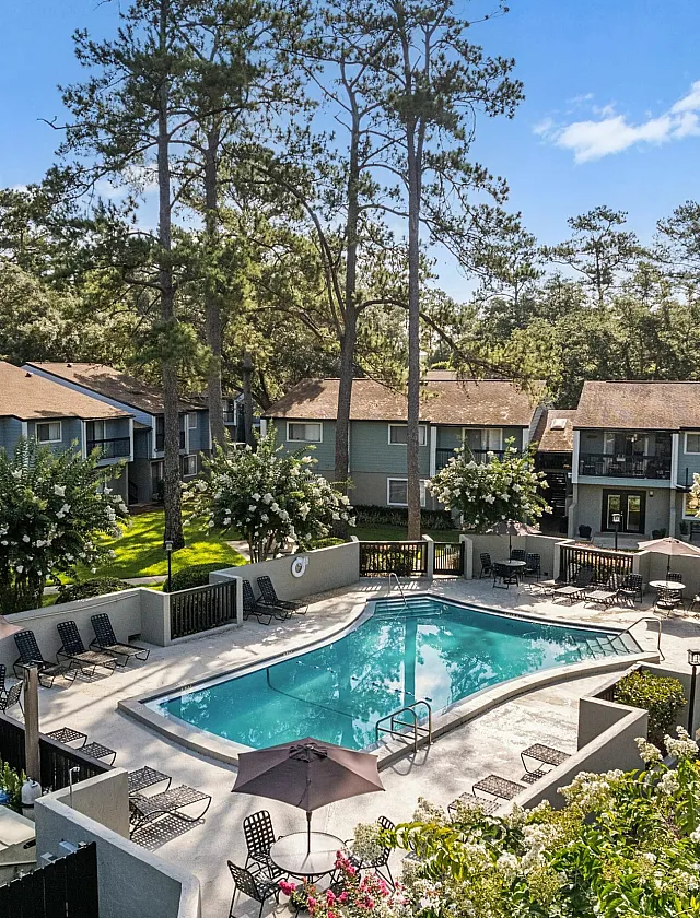 Charming apartment complex with lush greenery, a sparkling swimming pool, and inviting outdoor seating area under clear blue skies.