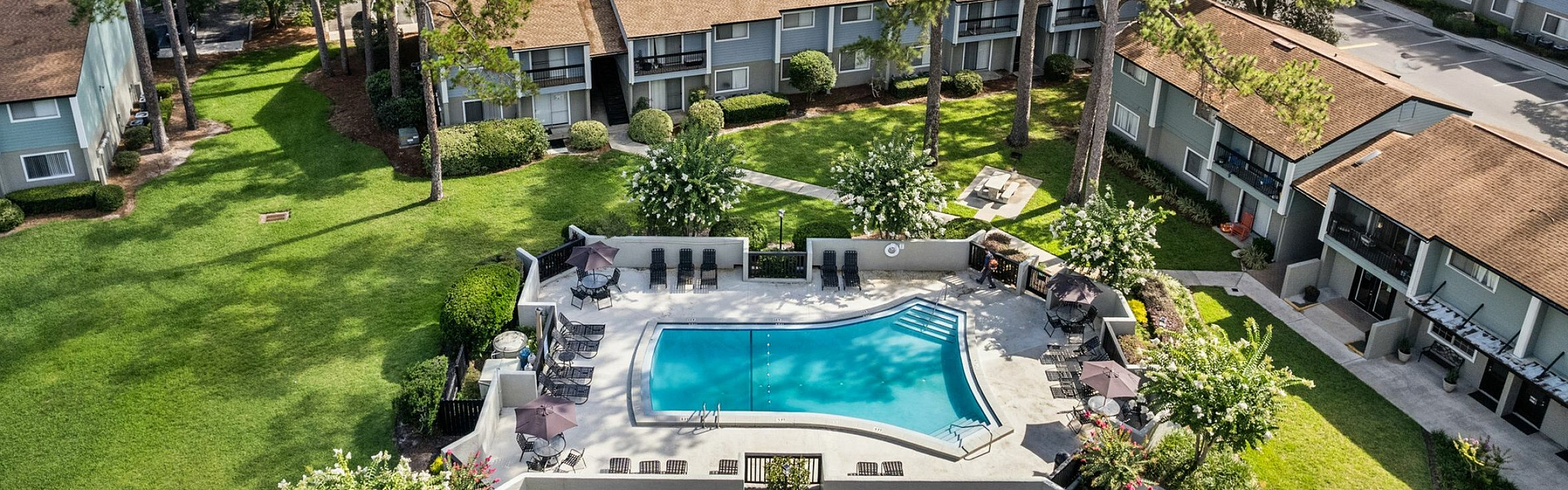 Aerial view of a tranquil apartment community surrounded by lush greenery, featuring a sparkling swimming pool and cosy apartment units with private balconies.