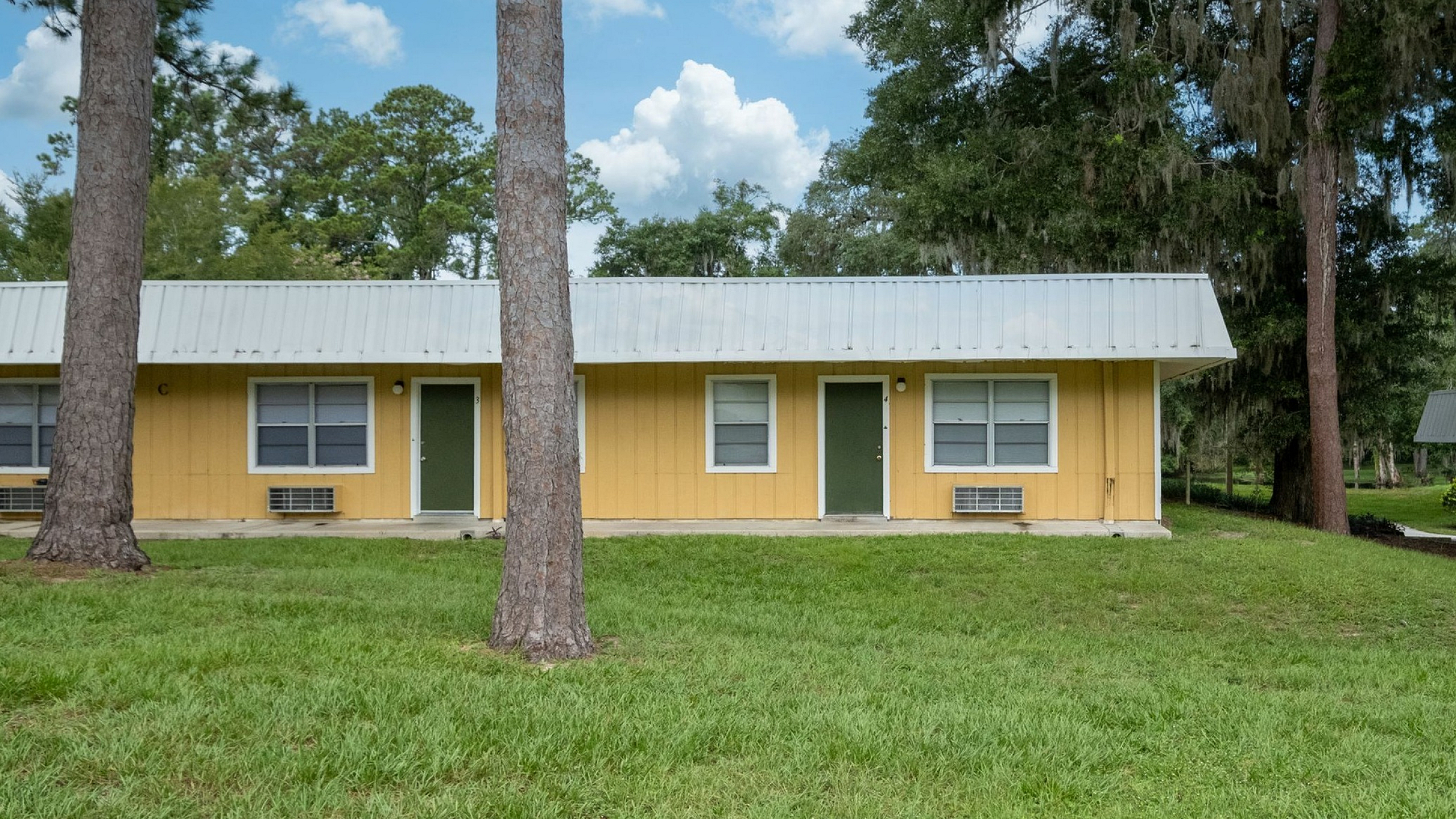 Charming single-story yellow home nestled among lush green trees, featuring a metal roof and spacious front lawn.