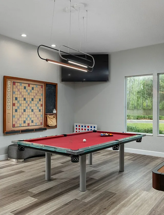 Contemporary recreation room featuring a red billiard table, modern lighting, and large windows overlooking lush greenery.