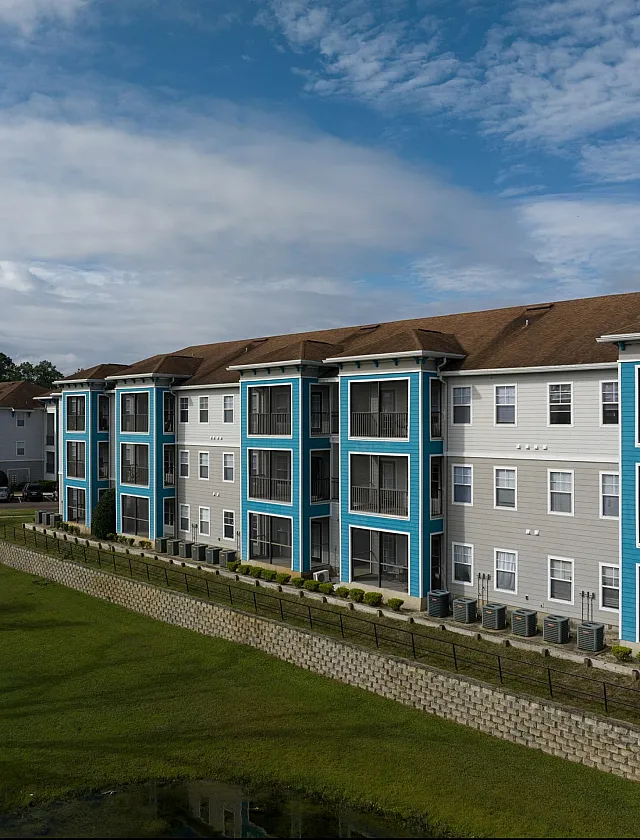 Modern apartment complex with vibrant blue accents, surrounded by lush greenery and a tranquil pond, set against a bright blue sky.