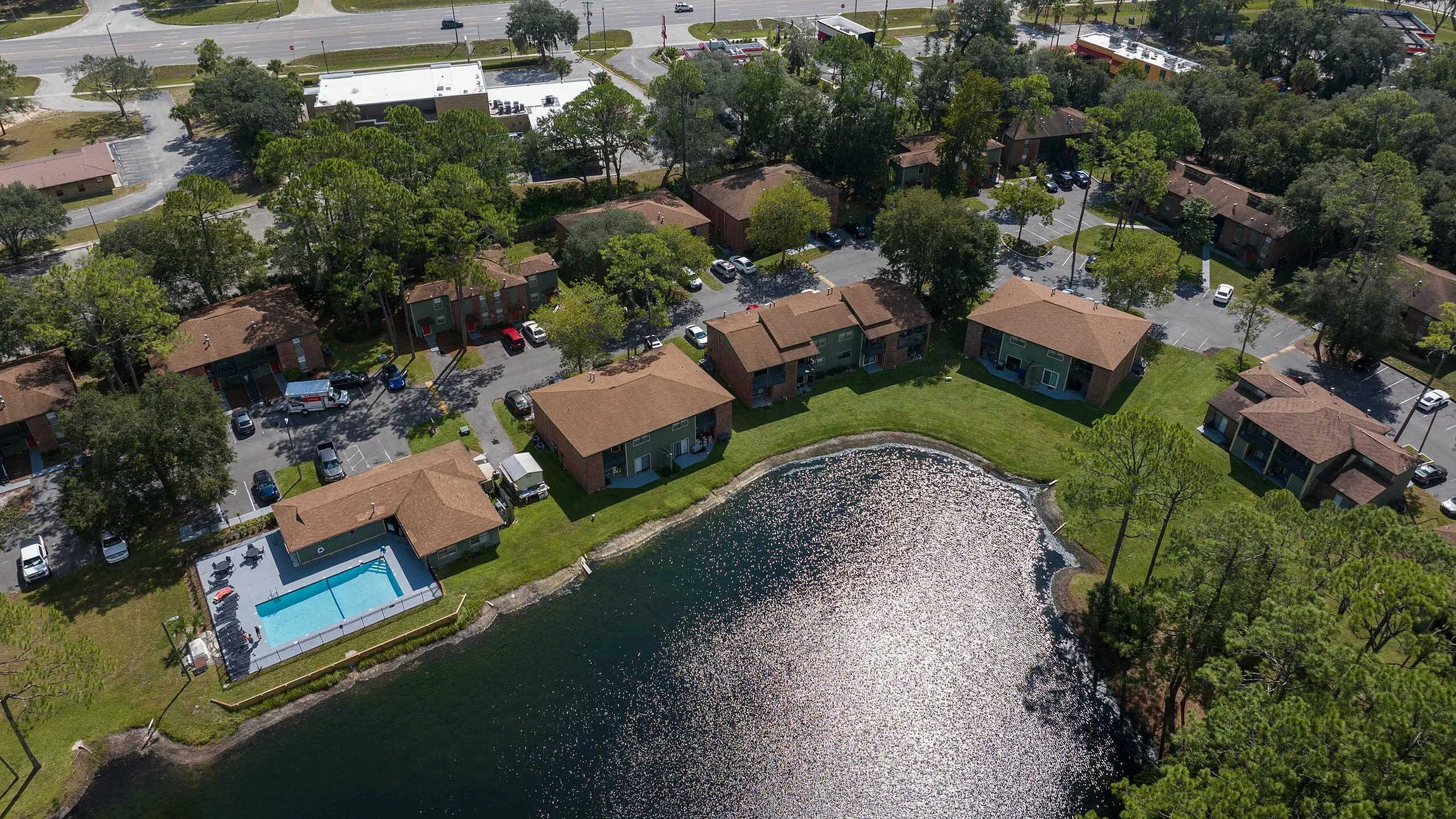 Scenic aerial view of a serene apartment complex nestled by a sparkling lake, featuring a refreshing swimming pool, lush greenery, and convenient proximity to urban amenities.